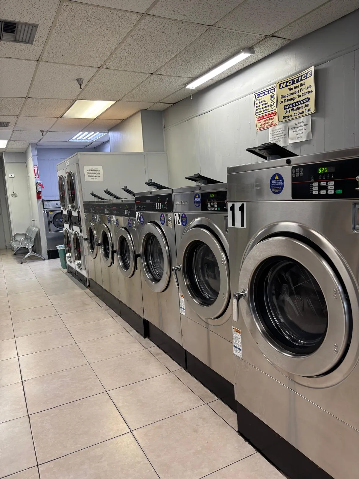 A bright tiled aisle inside the laundromat lined with large stainless steel Speed Queen front-load washers, numbered 11 and 12 in the foreground, with seating at the far end.