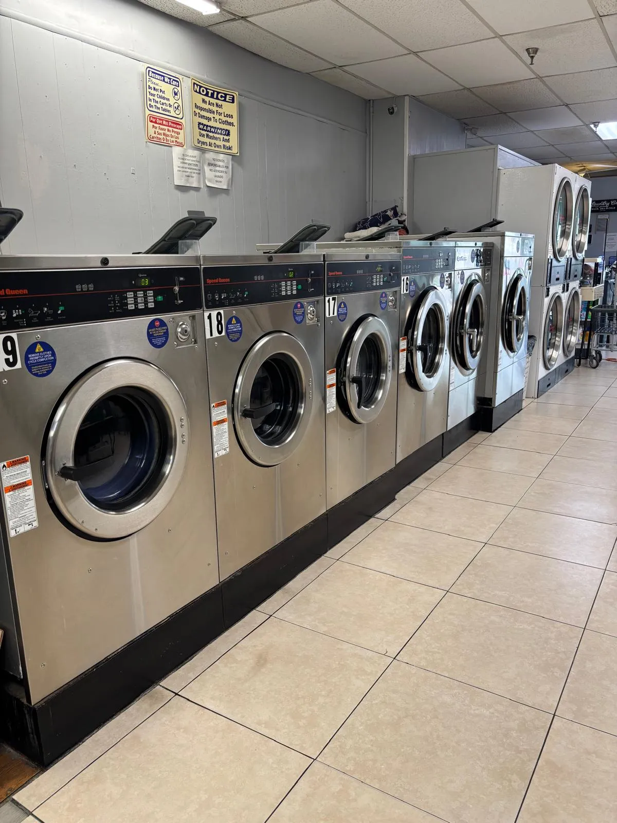 A row of stainless steel Speed Queen front-load washers numbered 16 through 19 along a clean tiled floor, with stacked dryers and the folding area at the back of the store.